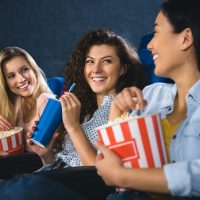 happy multiracial women with popcorn watching film together in movie theater